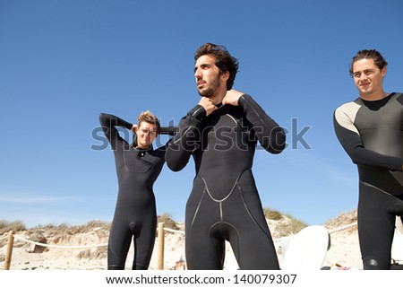 Three men friends getting dressed and ready with their surfing neoprene waterproof suits on a white sand beach with dunes and an intense blue sky in the background during a sunny day.