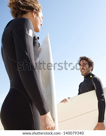 Two young surfers sports men wearing surfing neoprene waterproof suits and carrying their surfing boards on a white sand beach with a blue sky in the background during a sunny day.