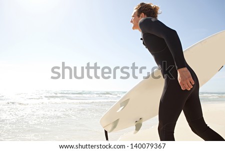Young surfer sport man carrying his surfing board and running into the sea  while on vacation during a sunny day, with an intense blue sky and light reflections in the water.