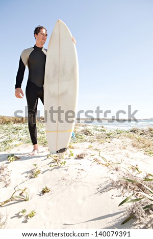 Young attractive surfer sports man wearing a neoprene rubber diving suit and looking at the horizon while standing on a beach during a sunny day with blue sky.