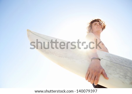 Young Attractive Surfer Sports Man Wearing A Neoprene Rubber Diving Suit And Looking At The Horizon While Carrying His Surfing Board During A Sunny Day Against The Intense Blue Sky.