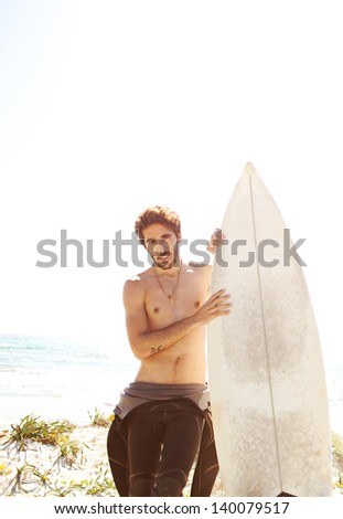 Young surfer standing on a white sand beach dunes, holding his surfing board and getting ready for surfing while wearing neoprene rubber suit during a sunny day.