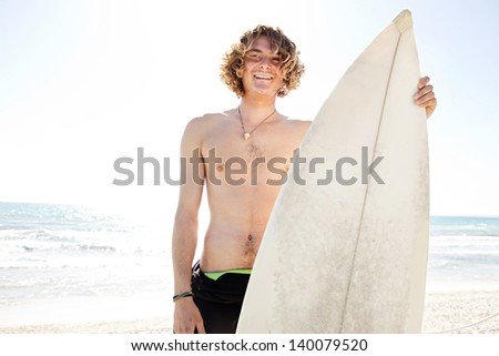 Portrait of a young surfer laughing while standing on a white sand beach with his surfing board against the blue sky and ocean on vacation.
