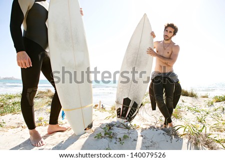 Two friends standing together on a white sand beach dunes, holding their surfing boards and getting ready for surfing while wearing neoprene rubber suits during a sunny day.