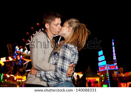 Young couple hugging and kissing while visiting an attractions park arcade at night, with colorful lights and rides around them.