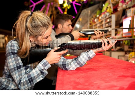 Young couple playing shooting games while visiting an amusement park arcade at night time, having fun with color lights and rides in the background.