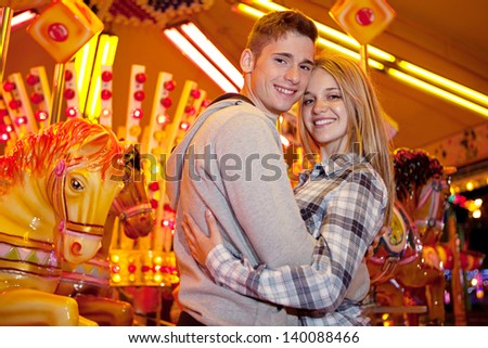 Young couple visiting an amusement park arcade together and hugging while standing next to a carousel ride with lights at night.