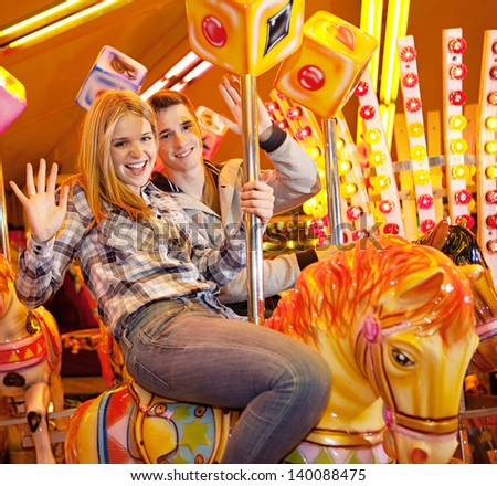 Young joyful couple visiting an attractions park and waving at camera while on a carousel ride during a fun night out together.