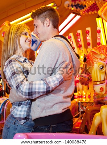 Young couple visiting an amusement park arcade together and hugging while standing next to a carousel ride with lights at night.
