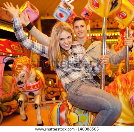 Young joyful couple visiting an attractions park and waving at camera while on a carousel ride during a fun night out together.