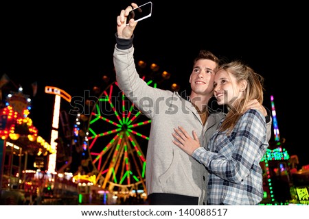 Teenager couple using a smartphone cell mobile to take a photograph while visiting an attractions park at night, with colorful lights and rides in the background.