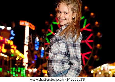 Portrait of a teenage girl visiting a funfair at night time during a night out, with colorful lights and rides in the background and a ferris wheel, smiling at camera.