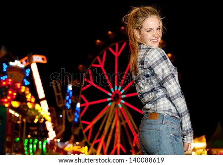 Portrait of a teenage girl visiting a funfair at night time during a night out, with colorful lights and rides in the background and a ferris wheel, smiling at camera.