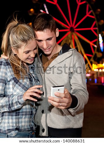 Teenager couple using a smartphone cell mobile while visiting an attractions park at night, with colorful lights and rides in the background.
