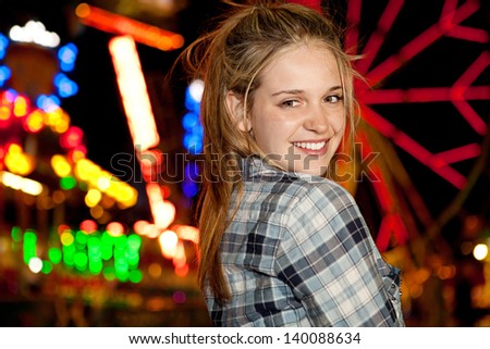 Close up portrait of a teenage girl visiting a funfair at night time during a night out, with colorful lights and rides in the background and a ferris wheel, smiling at camera.