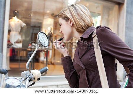 Young woman applying lipstick while in a shopping district street, using the reversing mirrors of her motorbike to put make up on.