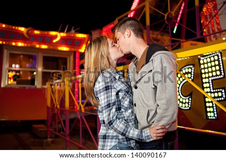 Side portrait of a young teenage couple visiting an attractions park arcade being romantic and kissing with funfair rides and lights in the background during night time.