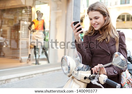 Young Woman Using A Smartphone While Standing By Her Motorbike In The Shopping District Of A City, With A Fashion Store Window With Manikins In The Background.