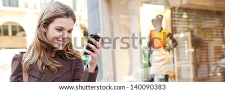 Young woman using a smartphone while standing by her motorbike in the shopping district of a city, with a fashion store window with manikins in the background.