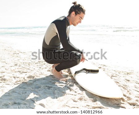 Young sport surfer man crouching next to his surfing board on a white sand beach while on a coastal vacation during a sunny day during the summer.