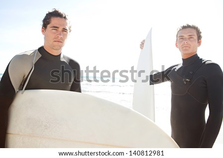Two surfers friends sharing a surfing trip experience, holding their surfing boards on a white sand beach with a sunny sky while on vacation and wearing specialist black neoprene suits.