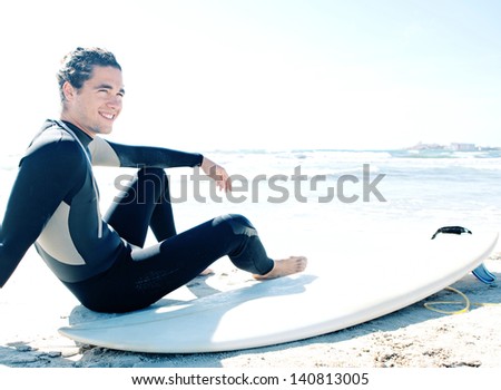 Young sport surfer man sitting down next to his surfing board on a white sand beach while on a coastal vacation during a sunny summer day, joyful and smiling.