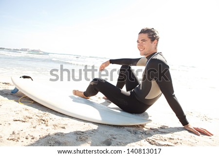 Young sport surfer man sitting down next to his surfing board on a white sand beach while on a coastal vacation during a sunny summer day, joyful and smiling.
