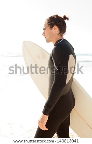 Young sport surfing man standing on a white sand beach contemplating the sea during a sunny day on holiday, being thoughtful and holding his surfing board.