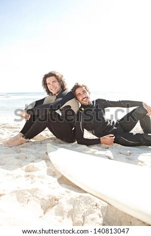 Two surfers friends sharing a surfing trip experience, laying down next to their surfing boards on a white sand beach with a sunny sky while on vacation and wearing specialist black neoprene suits.
