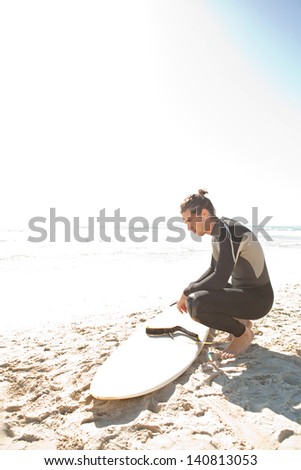 Side view of a young sport surfer man crouching next to his surfing board on a white sand beach while on a coastal vacation during a sunny day during the summer.
