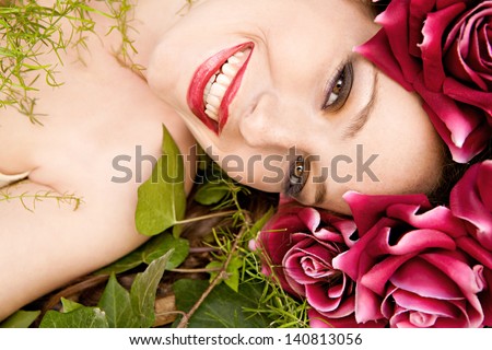 Close up beauty portrait of a young beautiful woman laying down in a forest wearing a red roses head dress hat while relaxing on a green bed of leaves and grass joyful and smiling.