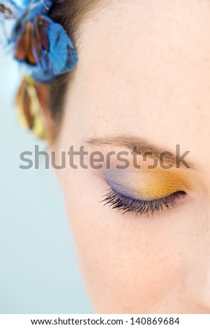 Close up portrait of a young woman half face wearing blue and yellow make up and a butterfly hair dress against a blue background with her eyes closed.