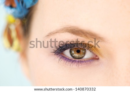 Close up beauty portrait of a young woman half face wearing blue and yellow make up and a butterfly hair dress against a blue background, intensely looking at camera.