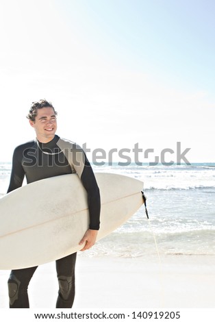 Surfer sports man during a surfing trip experience, holding his surfing board on a sunny white sand beach with a blue sky while on vacation and wearing a specialist black neoprene suit.