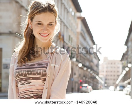 Young beautiful woman visiting a city center during a sunny day, smiling at the camera with buildings in the background.