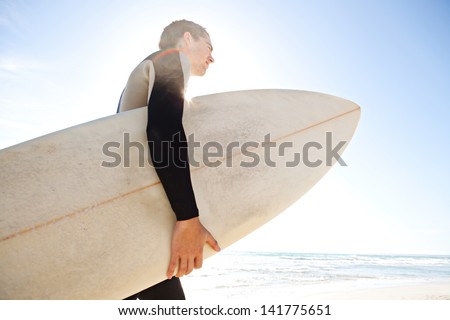 Profile view of a young surfer man carrying his surfing board under his arm and walking towards the sea with the sun filtering through his body during a sunny day with a blue sky.