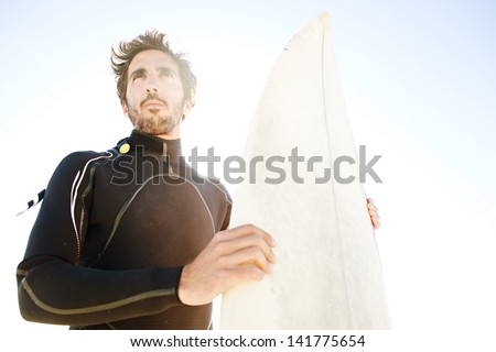 Close up aspirational portrait of an attractive young surfer holding his surfing board while being thoughtful against a sunny blue sky with the sun rays filtering through.