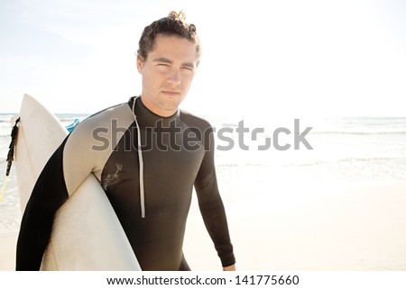 Young surfer man carrying his surfing board under his arm and walking away from the sea during a sunny day with a blue sky on vacation.