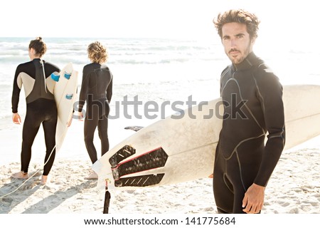 Three sport surfer friends standing together on a white sand beach carrying their surfing boards near the shore during a sunny golden day on vacation.