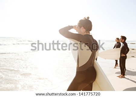 Three sport surfer friends standing together on a white sand beach carrying their surfing boards near the shore during a sunny golden day on vacation, looking out at sea.