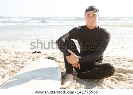 Young surfer man sitting down and relaxing on a white sand beach with his surfing board, wearing a black neoprene suit, rubber hat and goggles, during a sunny summer day.