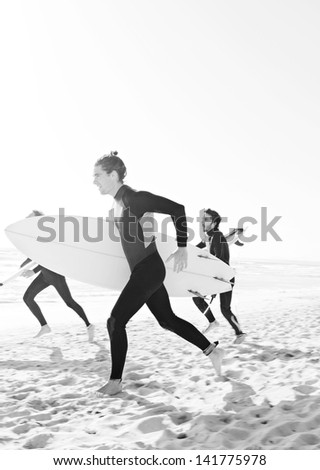 Black and white image of three sport surfer friends running together towards the sea on a white sand beach carrying their surfing boards during a sunny day on vacation.