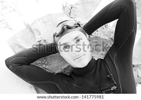Black and white over head portrait view of a young surfer man relaxing on a white sand beach with a surfing board, wearing professional neoprene, goggles and rubber hat, aspirational lifestyle.