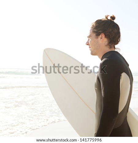 Side portrait of a sport surfer standing on a white sand beach carrying his surfing board under his arm on the sea shore during a sunny golden day on vacation, looking out at sea.
