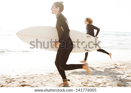 Two young surfers running together towards the ocean while carrying their surfing boards under their arms during a sunny day on a white sand beach.