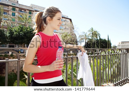 Side view portrait of a young woman taking a break from exercising in a city, holding a bottle of mineral water and smiling against a blue sky with buildings.