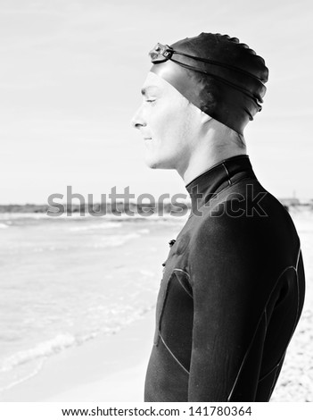 Black and white portrait of professional swimmer wearing a neoprene suit and a rubber hat getting ready for training, standing on a white sand beach during a sunny day.