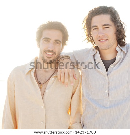 Two young men friends hanging around on vacation together with their arms around each other at sunset while on a beach having fun during the summer.