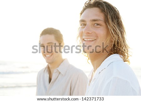 Portrait of two young men friends hanging around together on a golden beach on vacation, relaxing during a summer sunset and smiling at the camera.