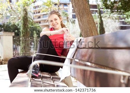 Sporty and attractive young woman sitting down on a wooden bench in the city, taking a break from working out and drinking mineral water during a sunny morning, smiling.
